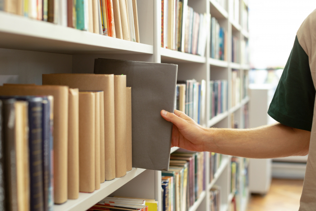 通信制大学の新入生が図書館で本を選ぶ写真、灰色の本を手に取る手と色とりどりの書棚が背景